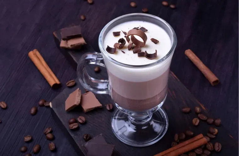 Un vaso con cafe en capas y crema decorada con rulos de chocolate, sobre una tabla rodeada de granos y canela.