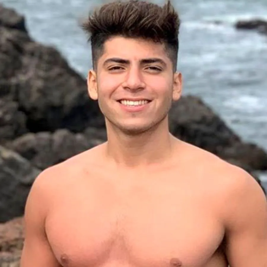 Joven sonriendo sin remera con el mar y rocas de fondo.