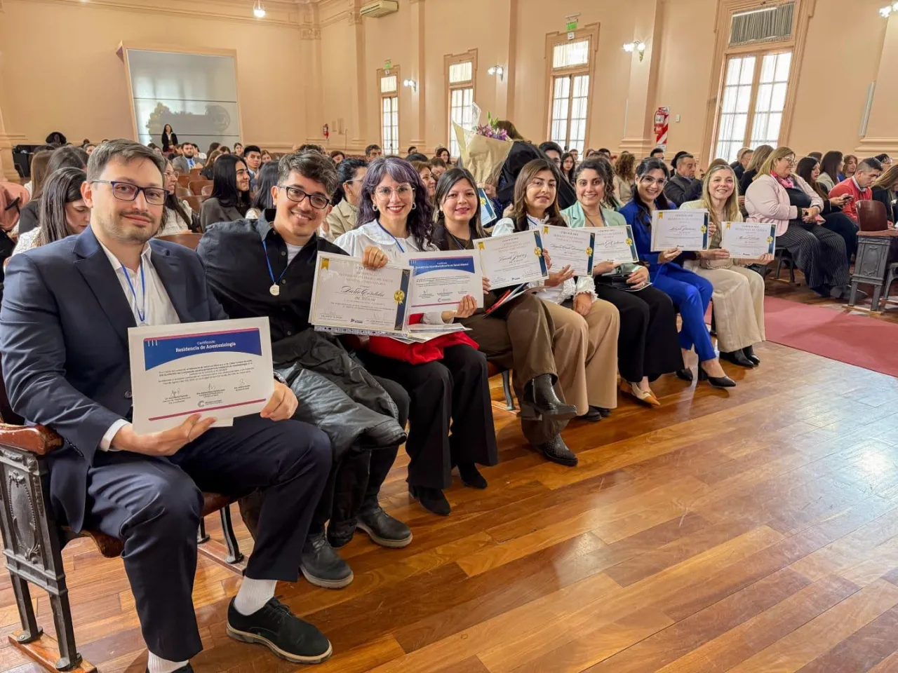 Un grupo de personas sentadas en una fila de butacas sostiene diplomas y certificados en un auditorio.