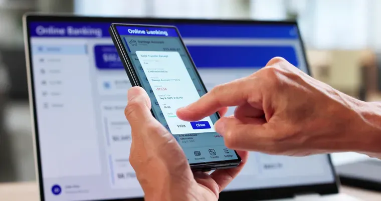 Una persona sostiene un teléfono celular mostrando un comprobante bancario frente a una computadora portátil.