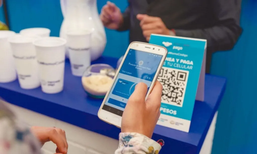 Una persona sostiene un celular frente a un cartel con código QR para pagar en un mostrador con vasos plásticos.