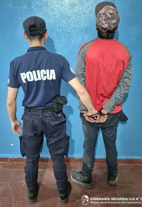Un policía de espaldas sosteniendo a un hombre esposado vestido con buzo rojo y gorra contra una pared celeste.