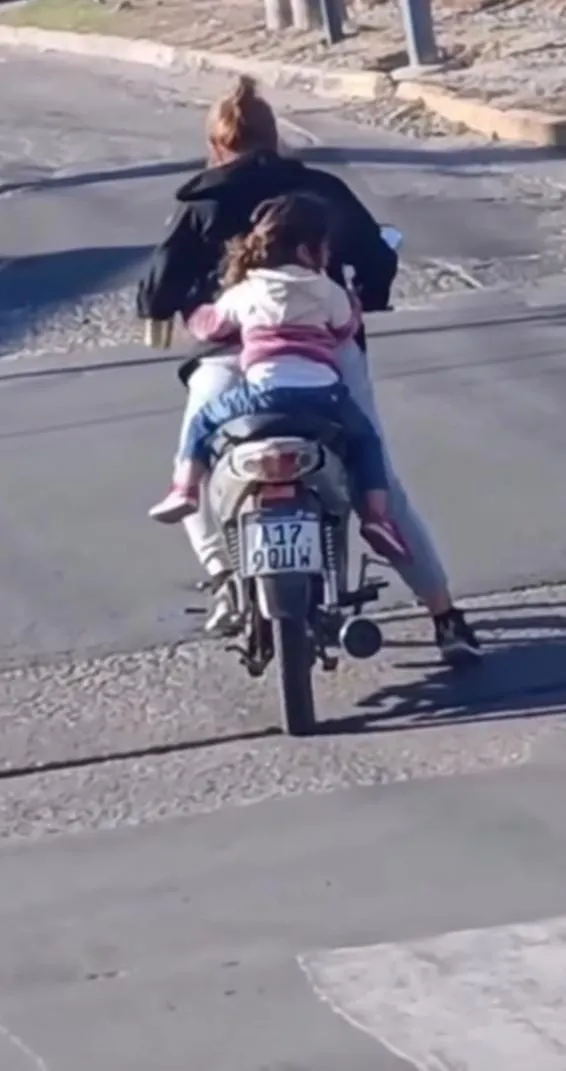 Una persona y una criatura viajan de espaldas en una moto por una calle asfaltada sin cascos de protección.