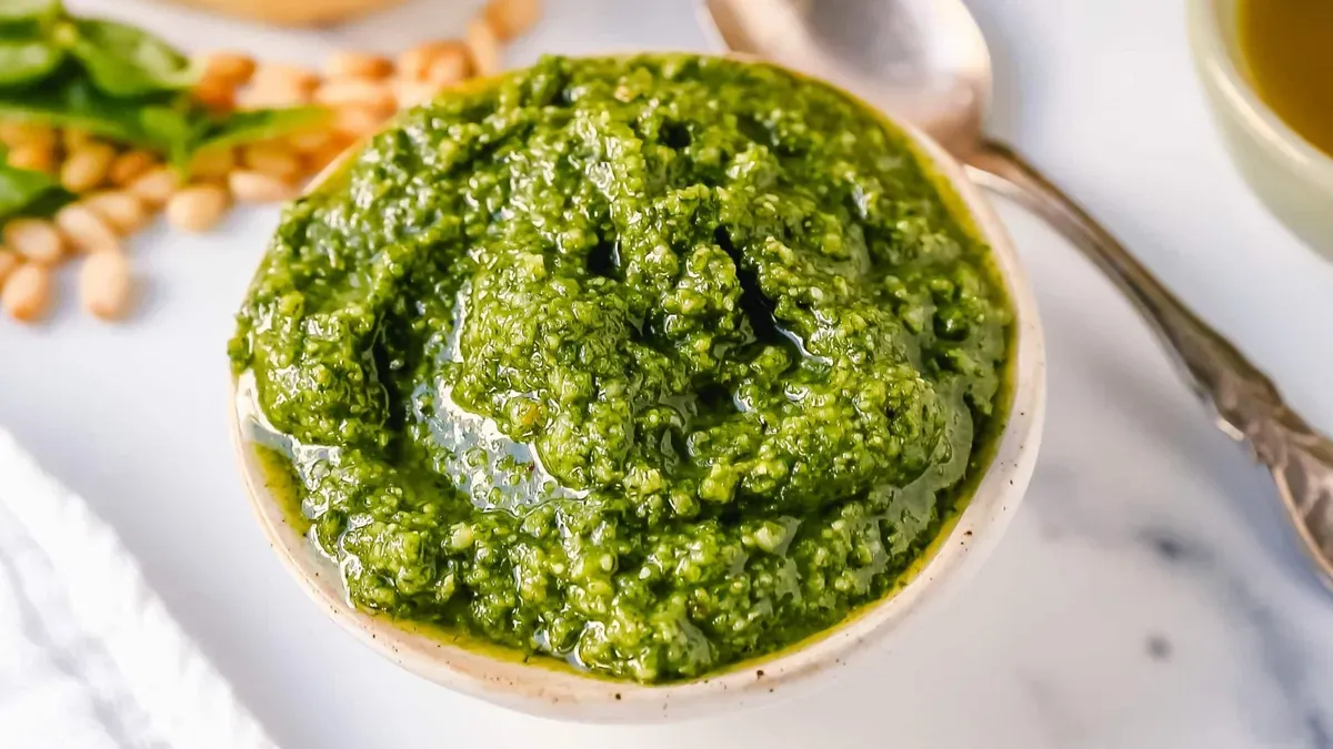 Un cuenco pequeño lleno de salsa pesto verde brillante junto a piñones y hojas de albahaca sobre la mesa.
