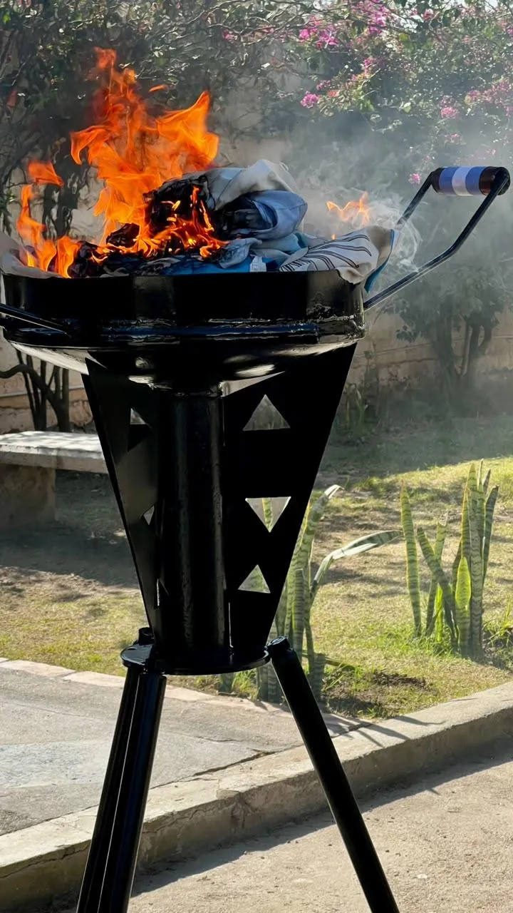 Prendas celestes y blancas arden dentro de un brasero metálico elevado sobre un trípode en un patio.