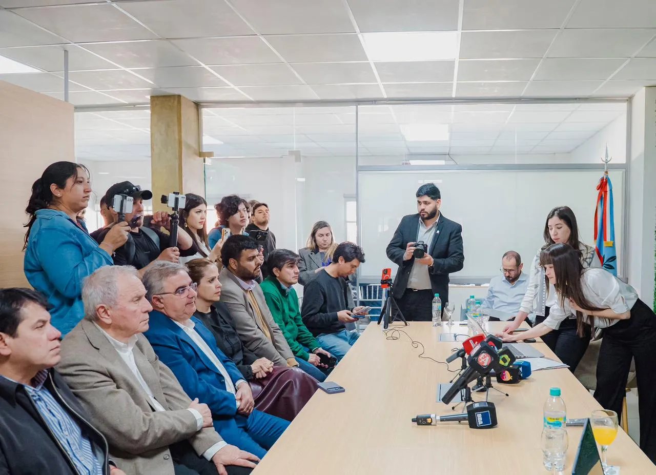 Un grupo de personas está reunido alrededor de una mesa con micrófonos mientras periodistas y camarógrafos registran el acto.