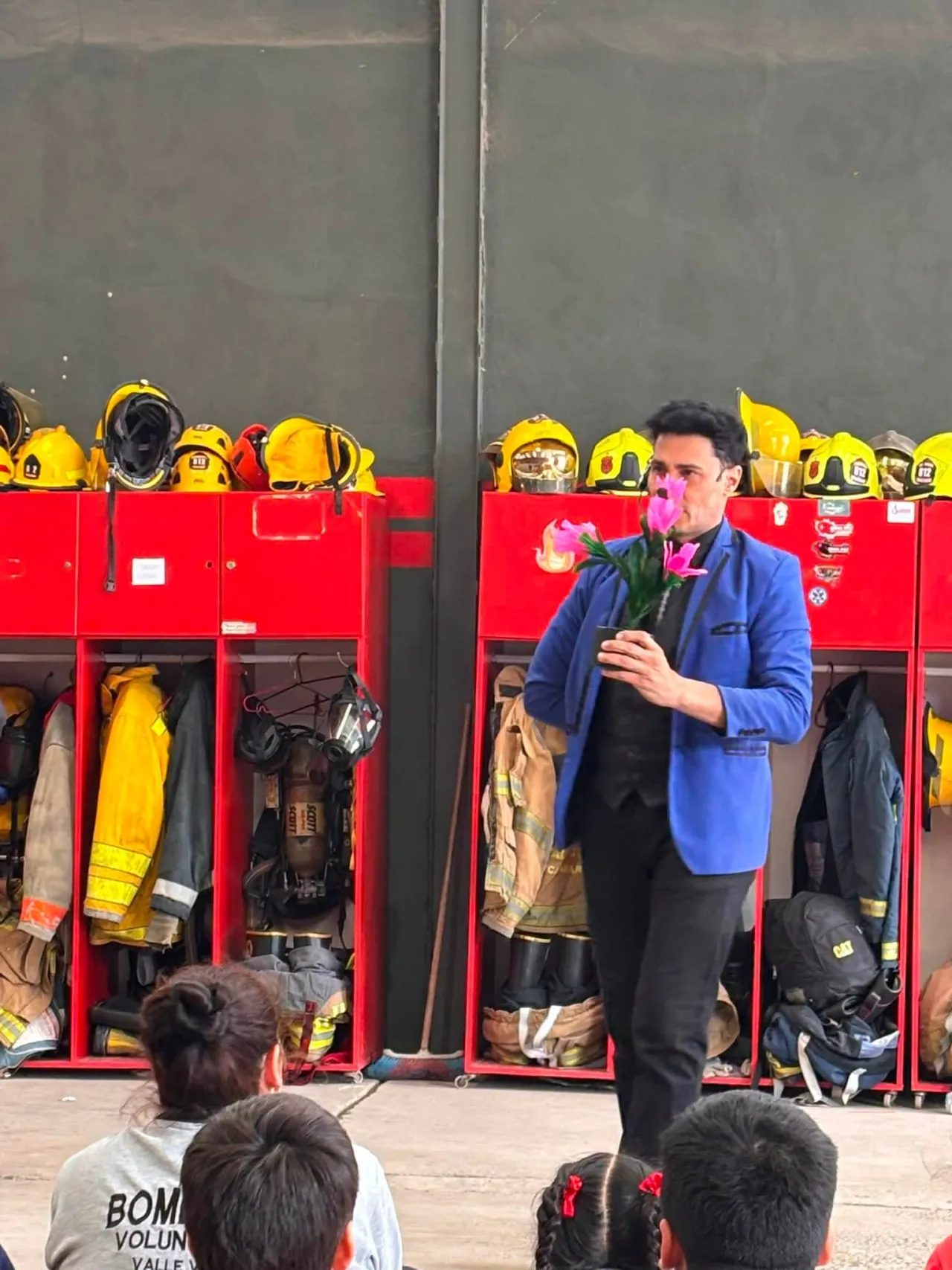 Un hombre de saco azul sostiene un ramo de flores rosadas frente a lockers rojos con equipamiento de bomberos.