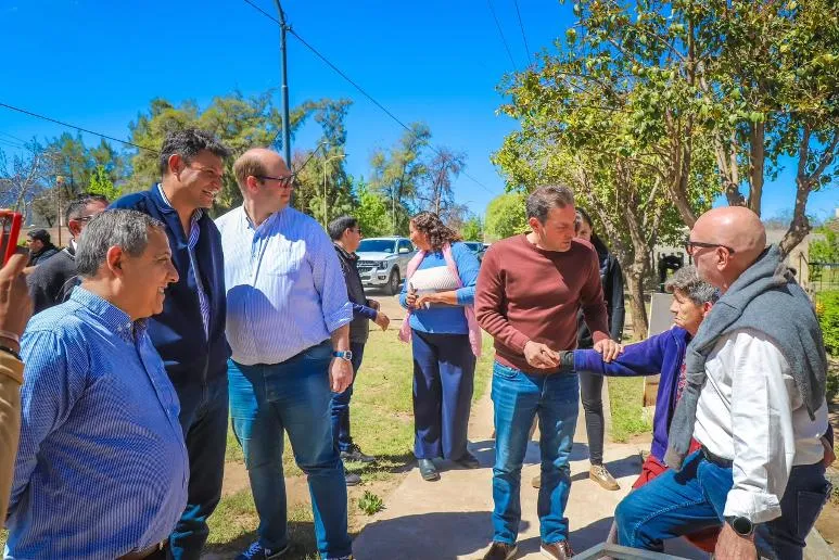 Varios adultos conversan al aire libre en un sendero rodeado de árboles y vegetación bajo el sol.
