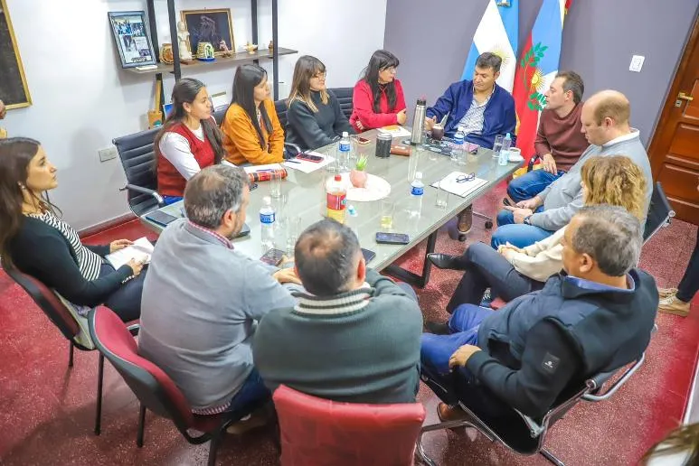 Un grupo de personas sentadas alrededor de una mesa de reuniones en una oficina con banderas de fondo.