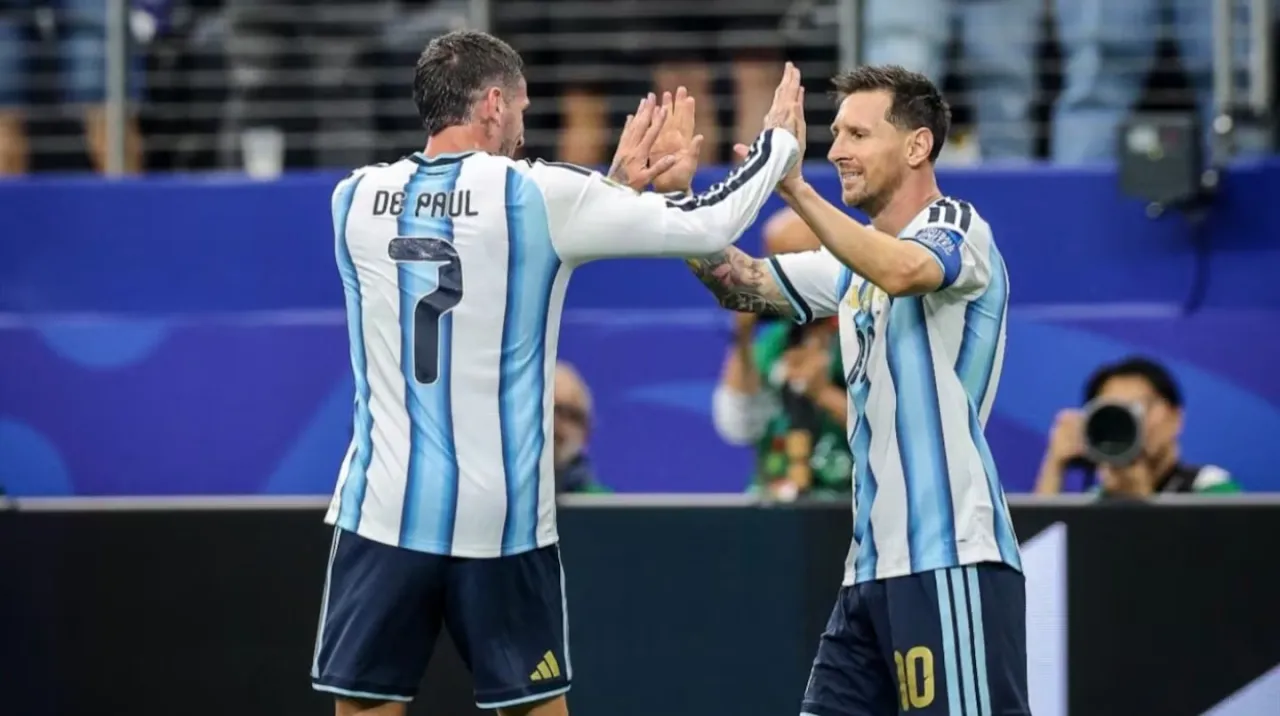 Rodrigo De Paul y Lionel Messi chocan las manos de espaldas y de frente en un campo de juego.