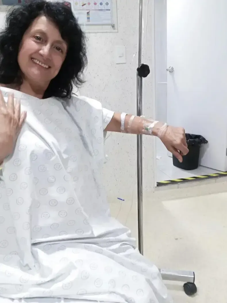 Una mujer con bata de hospital sonrie junto a un soporte con suero intravenoso.