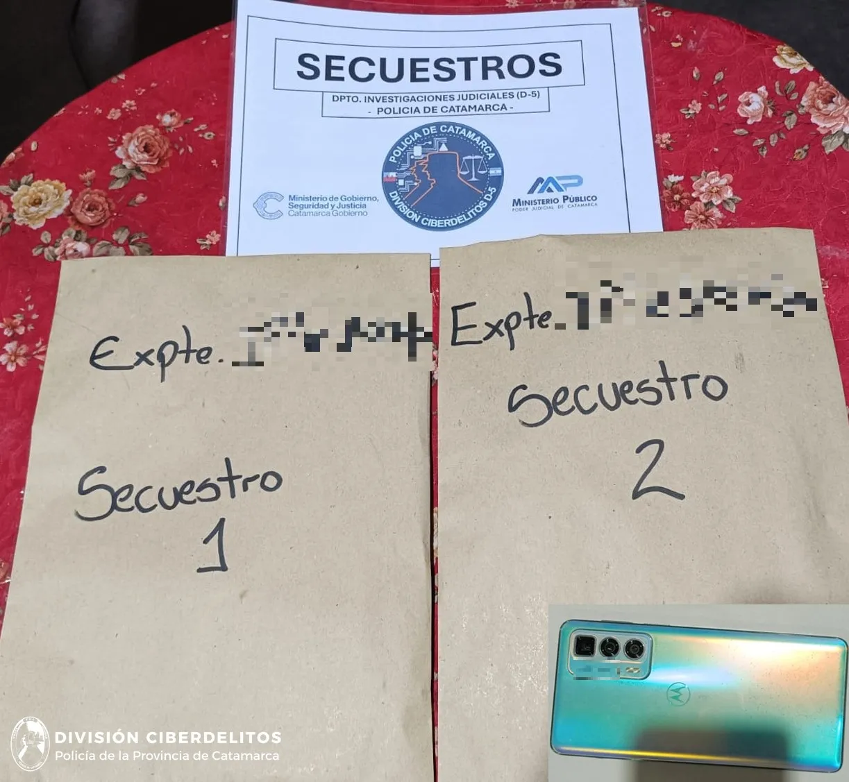 Dos sobres de papel madera rotulados, un cartel policial y un teléfono celular apoyados sobre un mantel floreado.