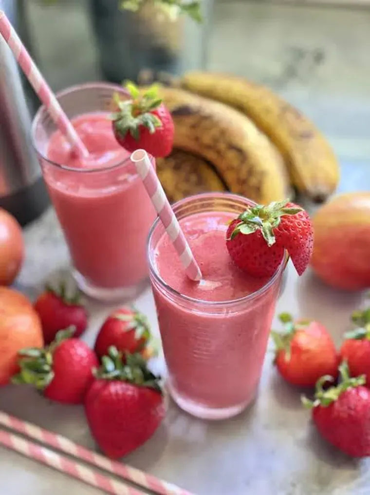 Dos vasos con licuado de frutilla con sorbetes junto a bananas y frutillas enteras sobre la mesa.