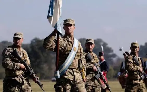 Soldados uniformados con fusiles desfilan al aire libre y uno de ellos lleva la bandera argentina.