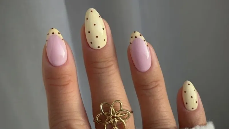 Los dedos de una mano muestran uñas esculpidas en tonos pastel con lunares y un anillo dorado con forma de flor.