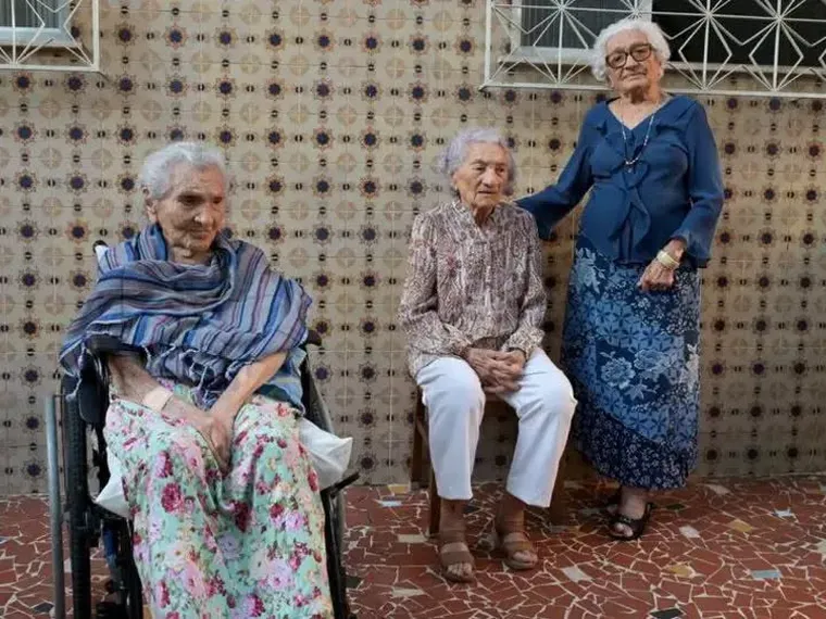 Tres mujeres ancianas posan juntas frente a una pared de azulejos, una de ellas sentada en silla de ruedas.