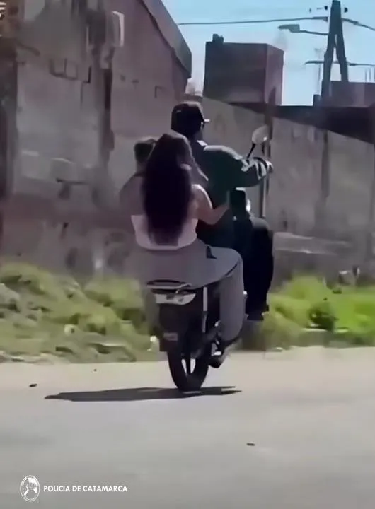 Tres personas van a bordo de una moto mientras el conductor hace willy por una calle.