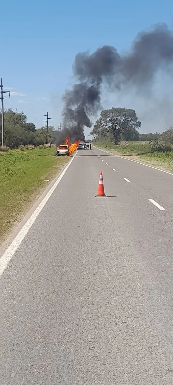 Un auto incendiándose en la banquina de la Ruta Nacional 64 con humo negro y un cono sobre el asfalto.