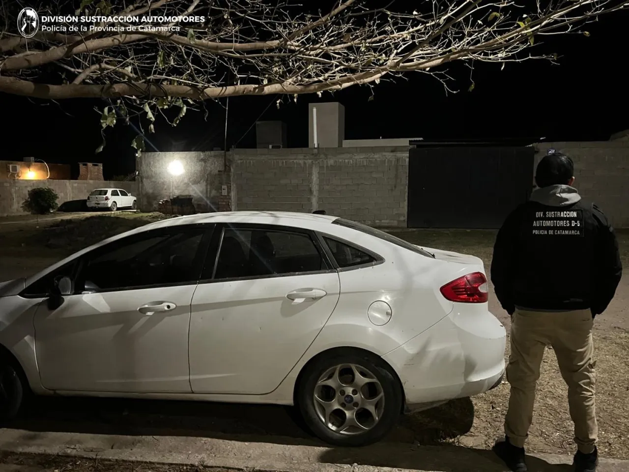 Un auto blanco estacionado de noche junto a un hombre de espaldas con una campera que dice Policía de Catamarca.