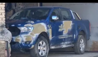 Una camioneta azul con masilla en su lateral y un hombre sentado al lado cerca del portón.