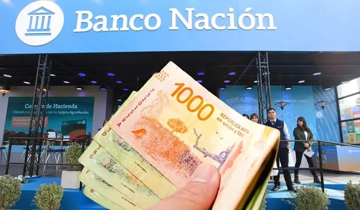 Una mano sostiene billetes de mil pesos argentinos frente a un cartel de Banco Nación.