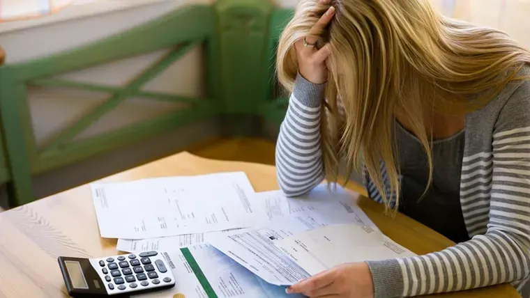Una mujer sentada a una mesa se toma la cabeza con una mano frente a facturas y una calculadora.