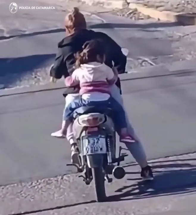 Una mujer y una nena van de espaldas en una moto por una calle asfaltada.