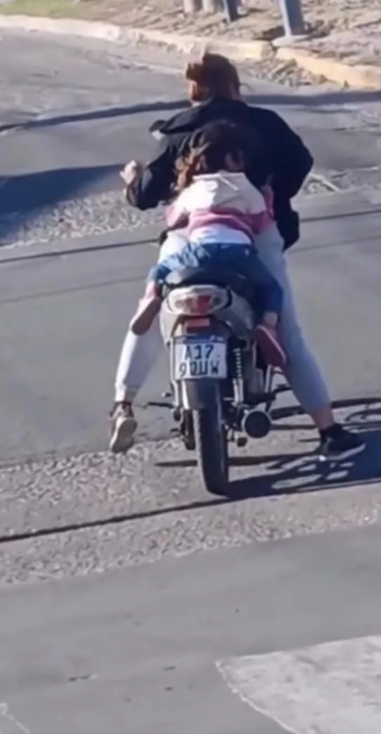 Una nena viaja sentada detrás de una persona en una moto detenida sobre el pavimento.