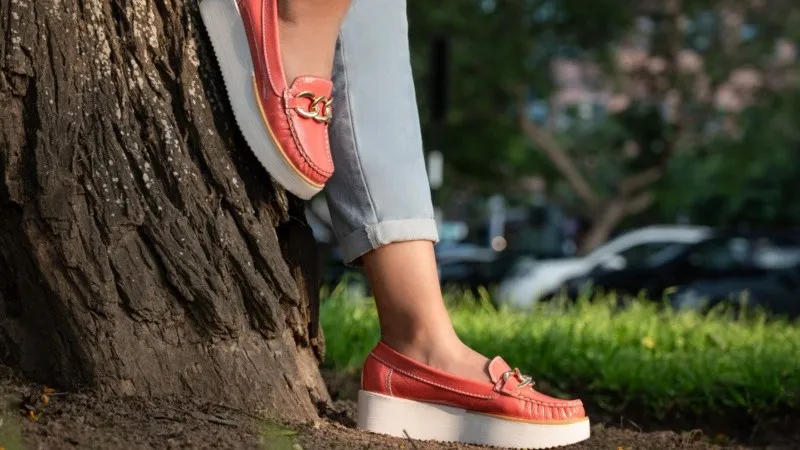 Una persona con mocasines rosas de plataforma y pantalón doblado se apoya contra el tronco de un árbol.