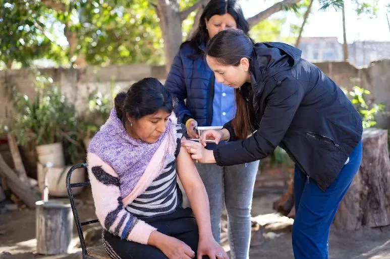 Una enfermera le aplica una inyección en el brazo a una mujer sentada al aire libre mientras otra persona observa.