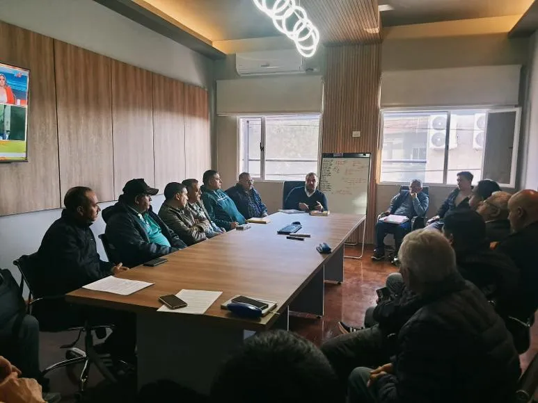 Un grupo de hombres sentados alrededor de una larga mesa de reuniones en una oficina con un televisor encendido.