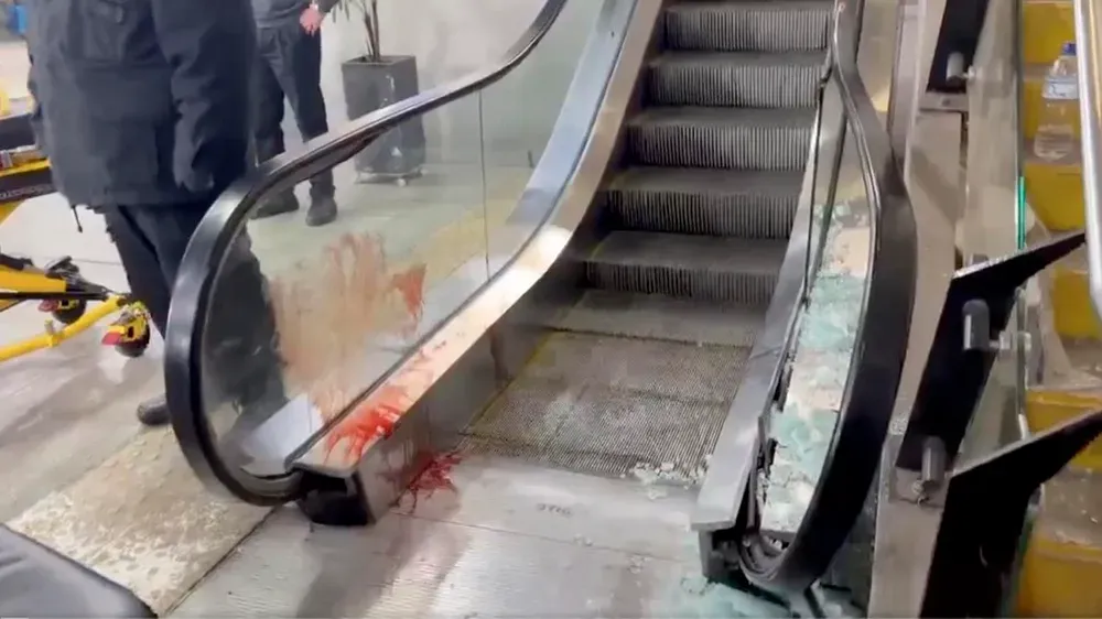 Una escalera mecánica con el vidrio lateral roto y manchas de sangre en la baranda y en los escalones.