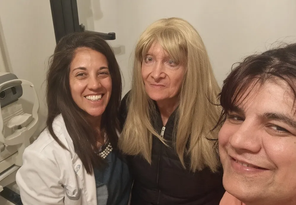 Una mujer con guardapolvo blanco y dos personas sonrien juntas mirando a la camara en un consultorio.