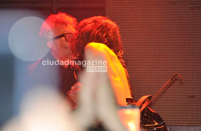 Fito Páez y una mujer se abrazan y besan en un evento bajo una iluminación tenue rojiza.