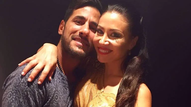 Un hombre y una mujer sonriendo abrazados frente a la camara sobre un fondo oscuro.