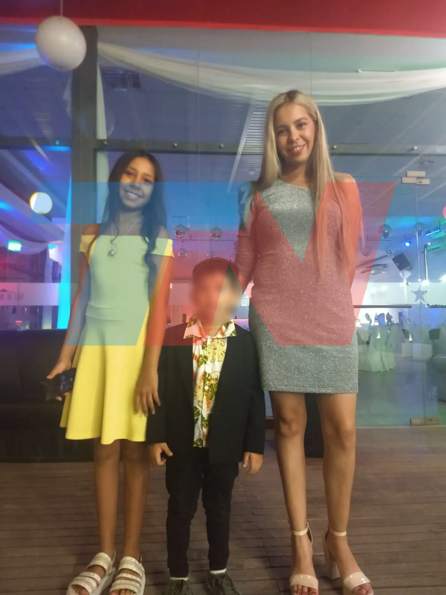 Una nena con vestido amarillo, un nene de saco con rostro borroso y una mujer con vestido plateado posan en un salon.