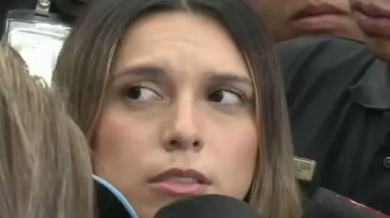 Una mujer de pelo castaño mira hacia un costado rodeada por personas y micrófonos.