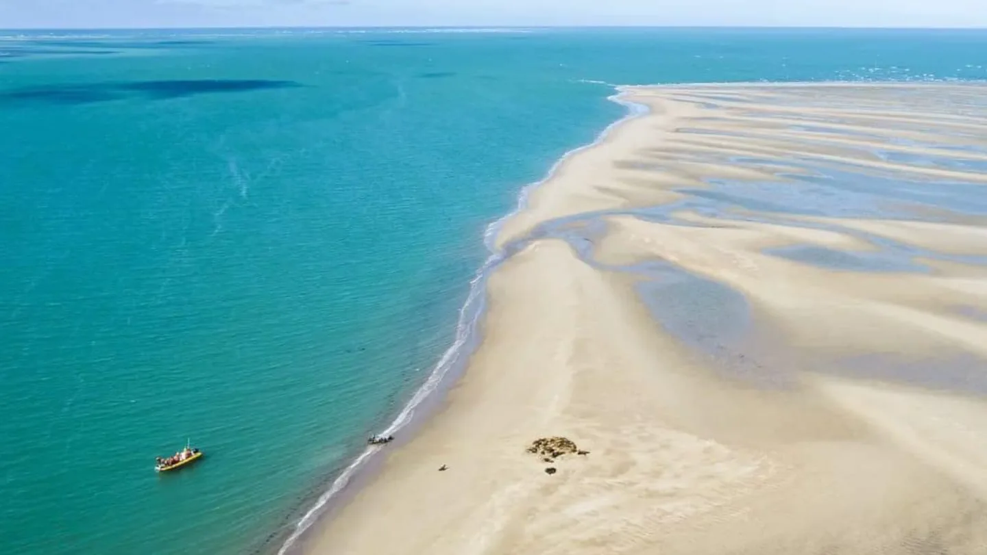 Vista aérea de un extenso banco de arena clara rodeado por aguas calmas de color turquesa con una lancha en la orilla.