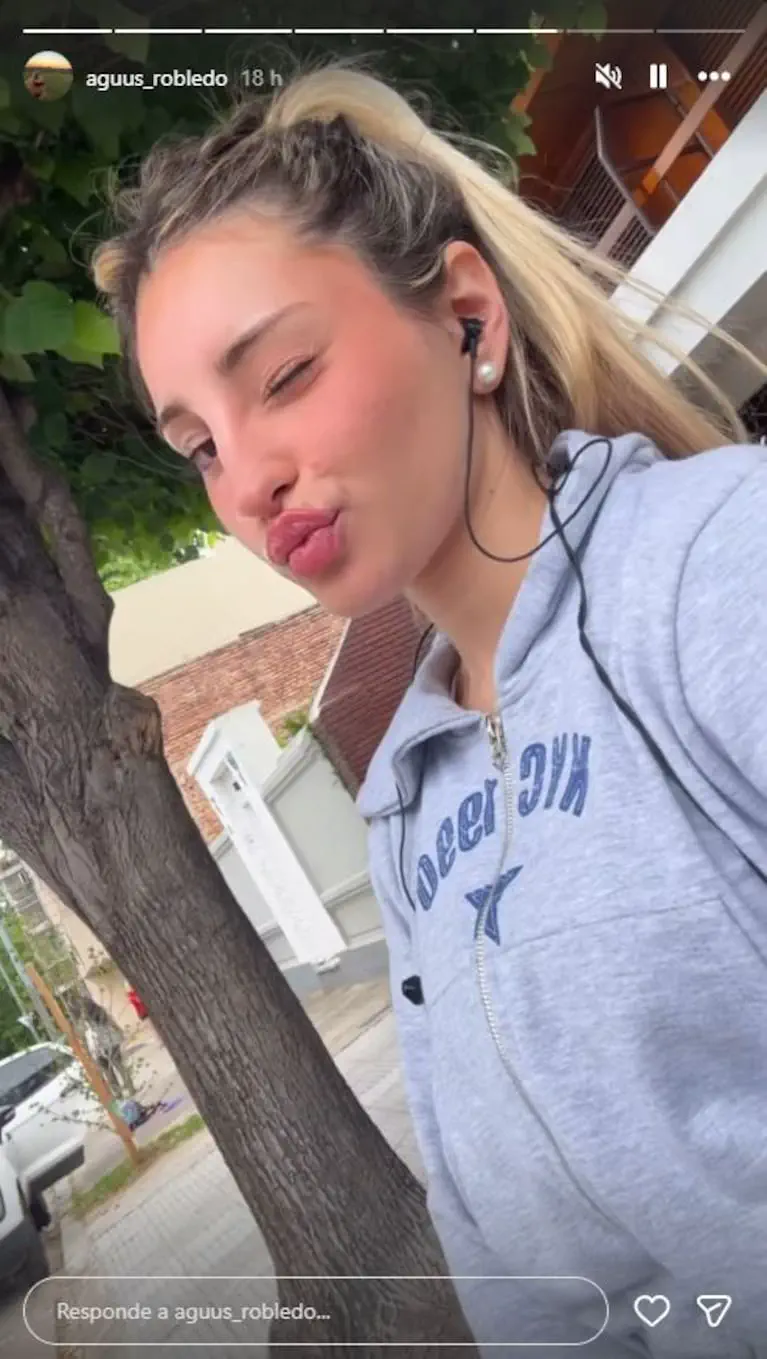 Una joven con campera gris y auriculares guiña un ojo e infla los labios al aire libre.