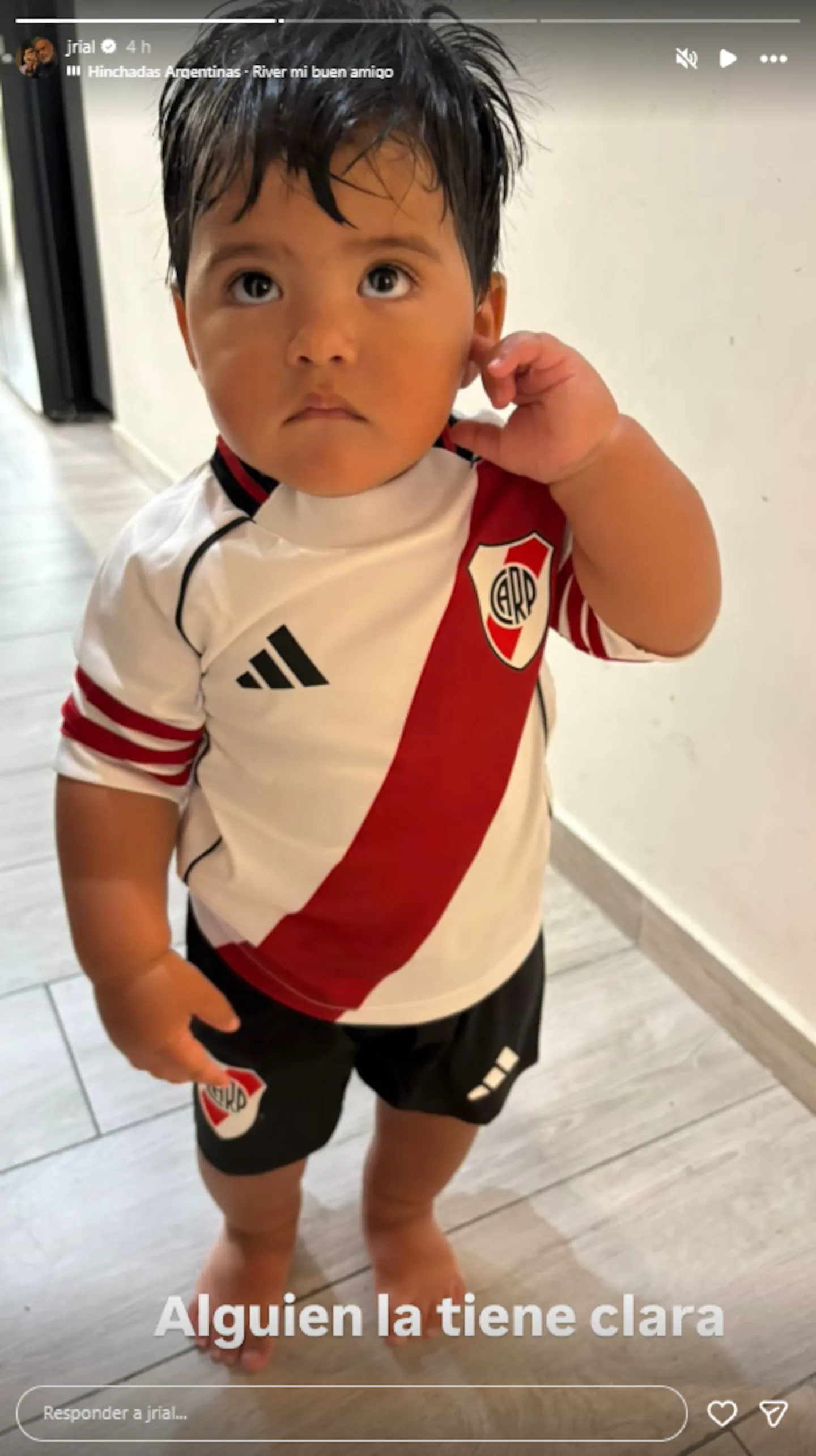 Un nene descalzo de pie con la camiseta y el pantalón corto del Club Atlético River Plate.