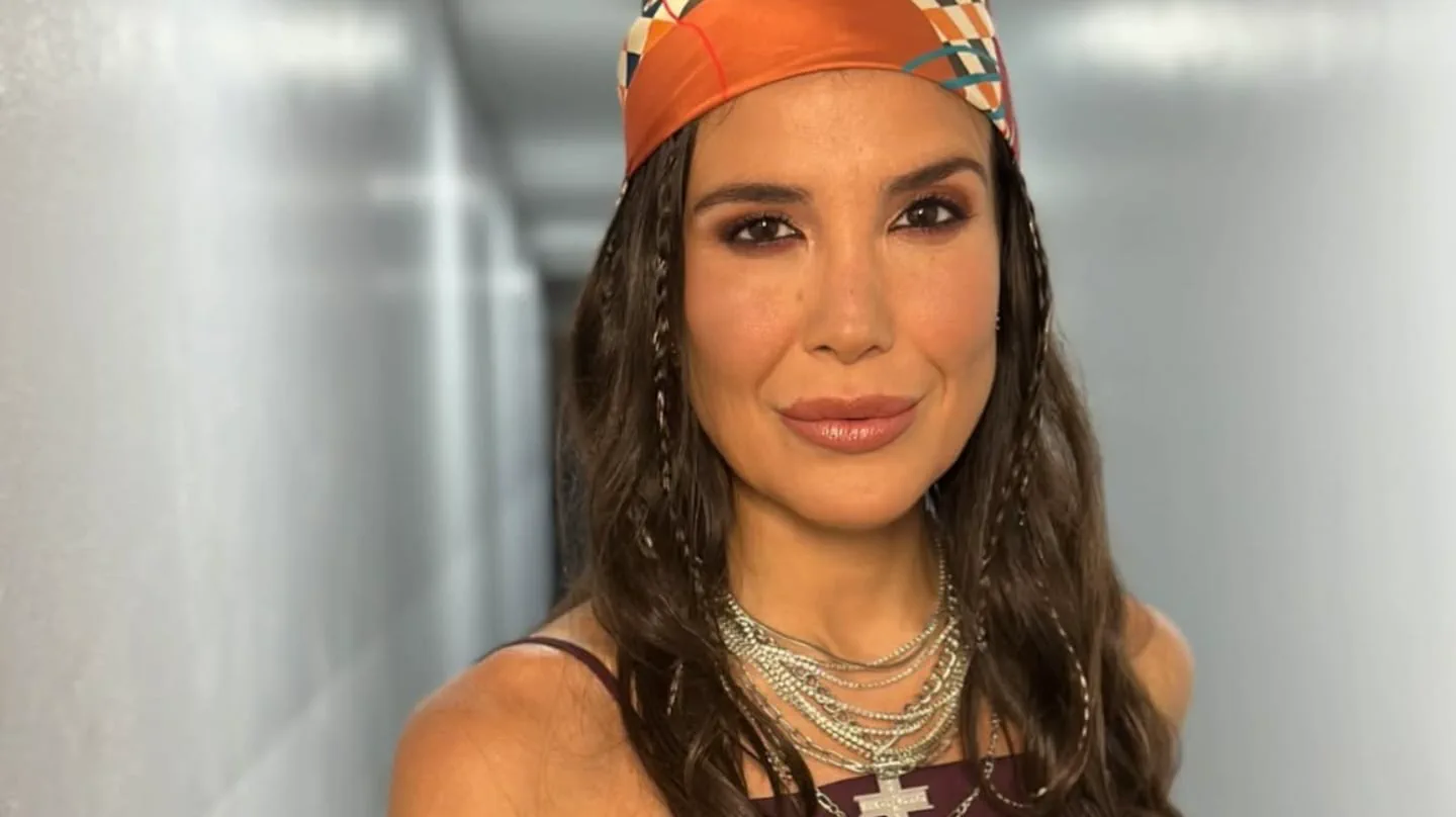 Una mujer con pañuelo naranja en la cabeza y collares plateados mira a la cámara en un pasillo.