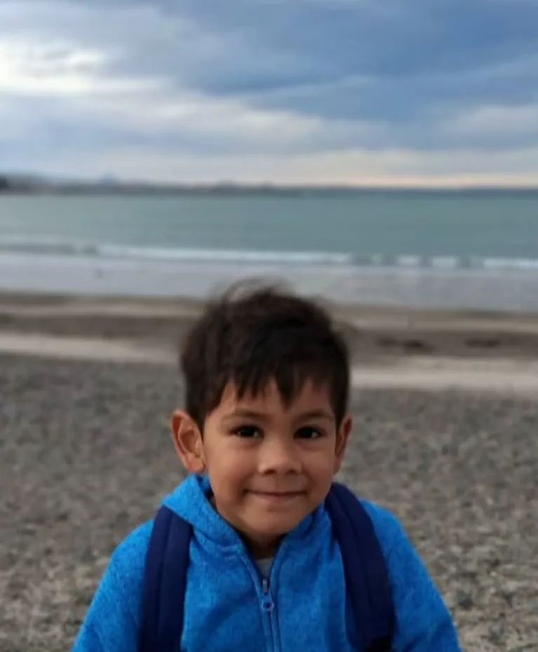 Un nene sonriente con campera azul posa en una playa de piedras frente al mar.