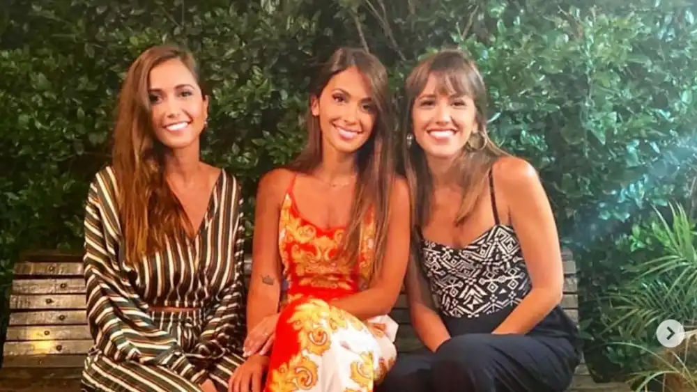 Tres mujeres sonriendo sentadas en un banco de madera con plantas de fondo, entre ellas Antonela Roccuzzo.