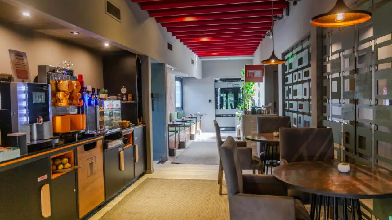 El salón de una cafetería o desayunador con mesas redondas, sillas tapizadas y máquinas de autoservicio.