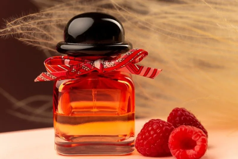 Un frasco de perfume rojo con un moño estampado descansa sobre una superficie clara junto a tres frambuesas.