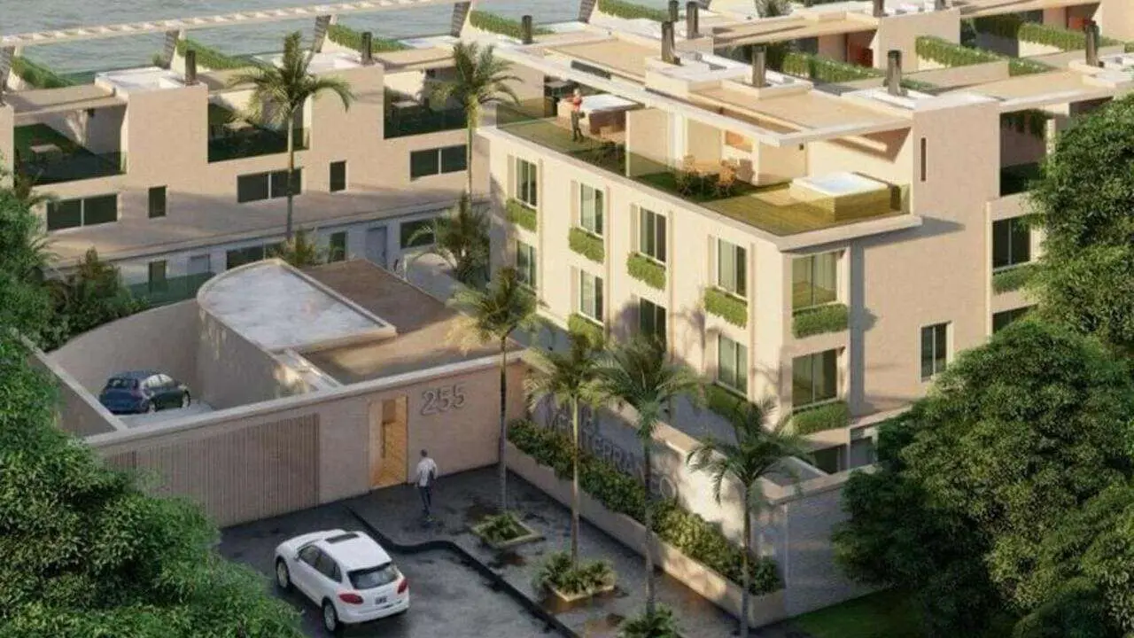 Vista aérea de un complejo residencial con terrazas, palmeras y autos estacionados en la entrada.