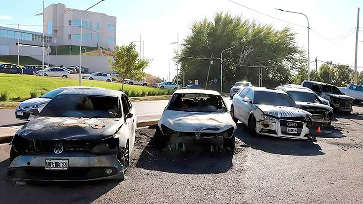 Varios autos quemados y destrozados se encuentran estacionados al costado de una calle frente a un edificio.