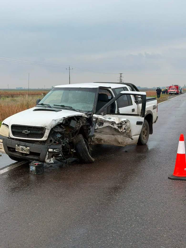 Una camioneta blanca chocada con graves daños en la parte delantera izquierda está parada sobre el asfalto mojado.