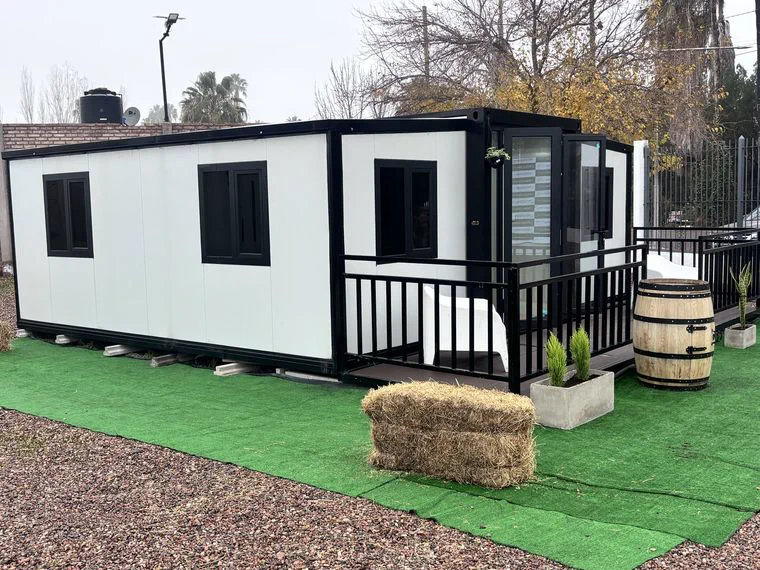 Una casa modular blanca y negra con baranda, fardo de pasto y barril sobre cesped sintetico.