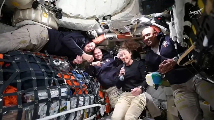 Cinco astronautas sonrien y posan flotando dentro de la estacion espacial mientras una mujer sostiene un microfono.